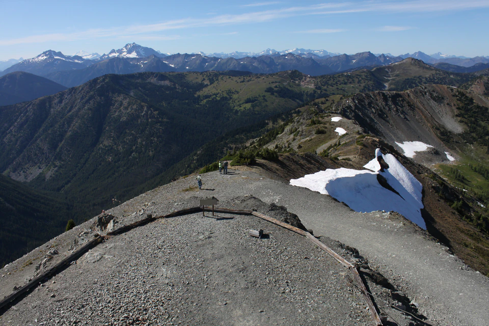 Rocky summit ridge and lingering snow below Slate Peak Lookout, with jagged North Cascade peaks on the horizon.